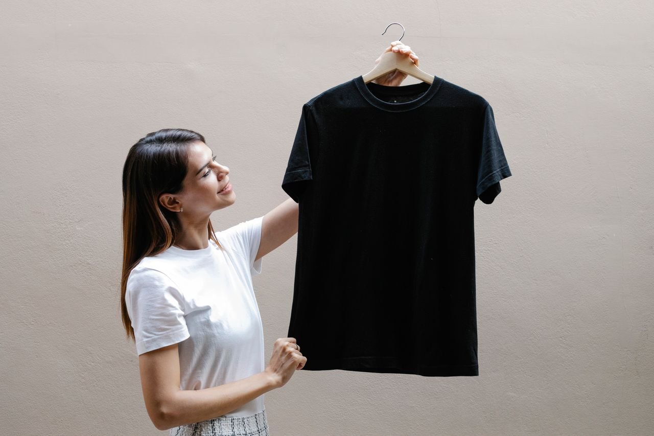 Home Side view of self assured young woman with long dark hair in stylish outfit smiling while holding hanger with black t shirt against gray wall