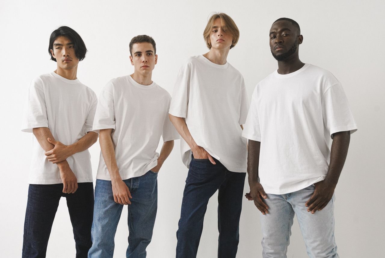 Home A group of four diverse men modeling white t-shirts and jeans in a studio setting.